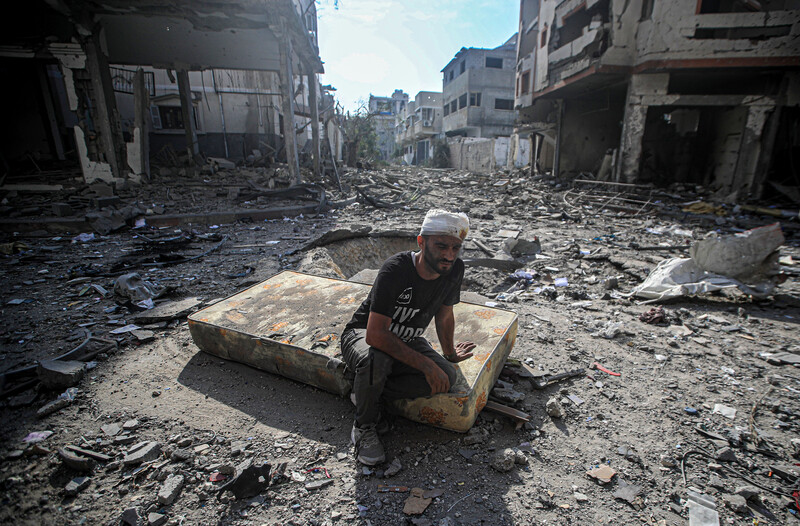 A man with a bandaged head sits on a mattress in a debris-filled street