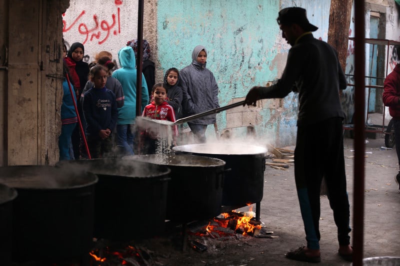 A man holds a ladle over a steaming vat of soup as people stand nearby