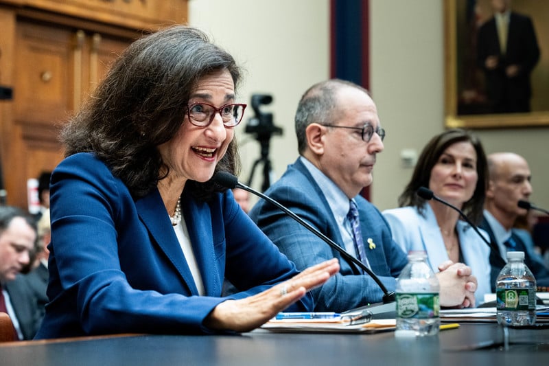 Minouche Shafik and two others sit at a table in a hearing room