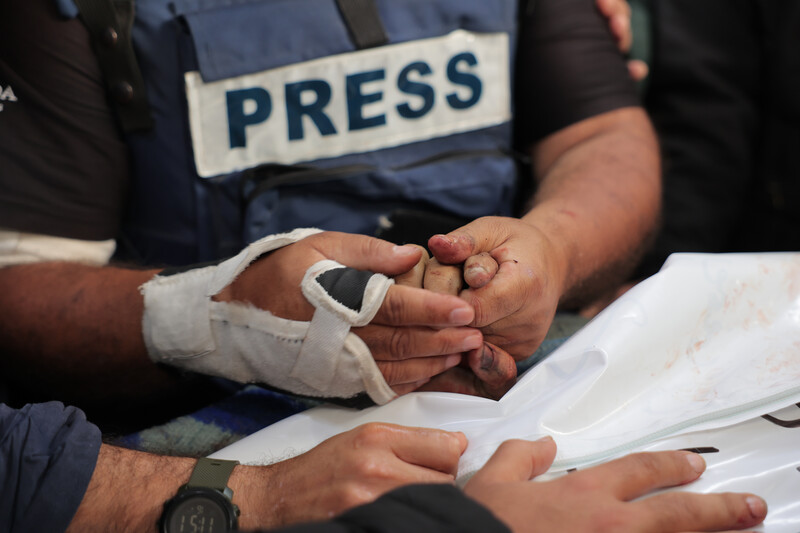 A man wearing a press vest holds the hand of someone wrapped in shroud