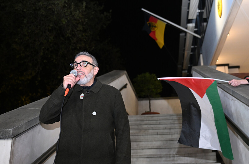 Dr. Abu Sitta speaks in a microphone outside a building with German and Palestinian flags visible behind