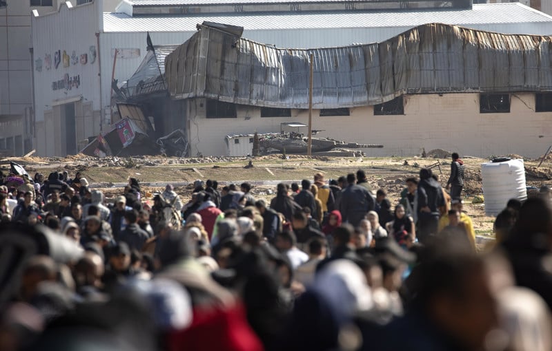 A crowd of people is overlooked by an armored tank in the background