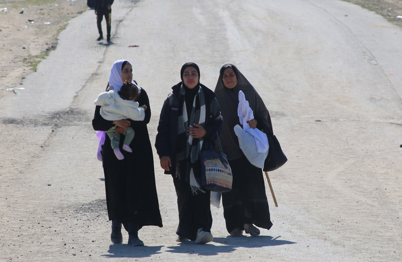 three women walk on a dusty road, one carrying an infant