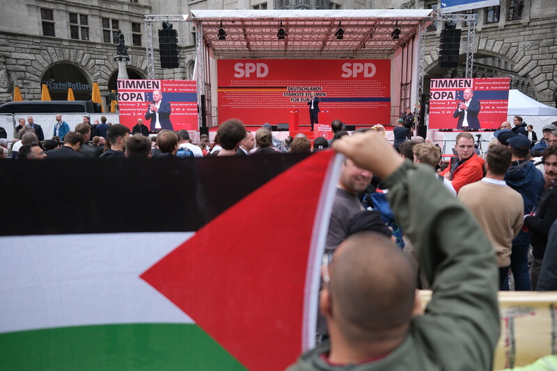 Man holds large Palestinian flag in foreground with man on stage in background