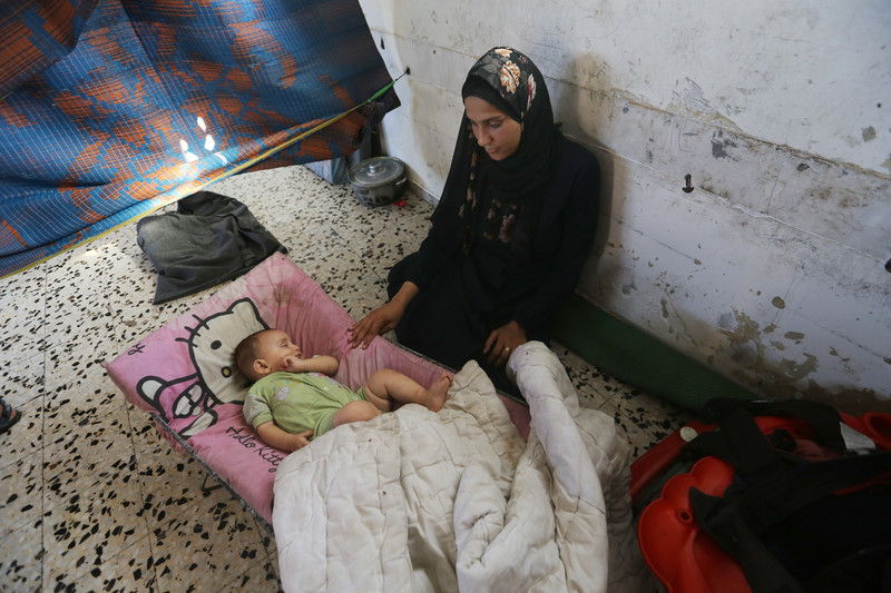 A woman sits against a wall, touching her sleeping infant. 