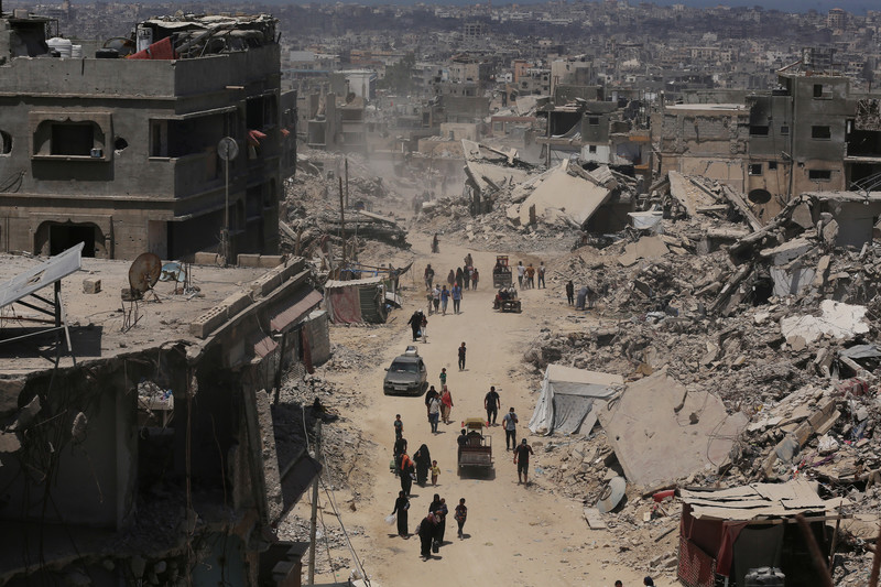 Aerial view of dozens of people walking along sandy road in between collapsed and bombed-out buildings