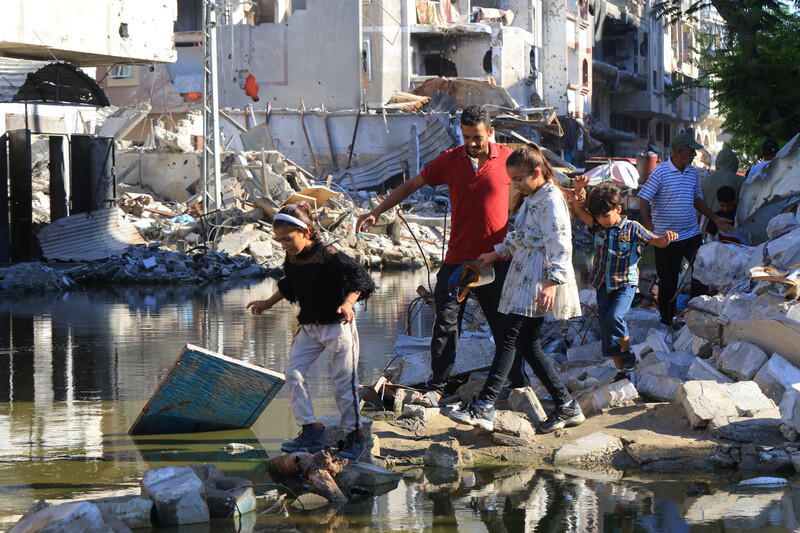 People walk through a pool of sewage water amid the rubble of buildings