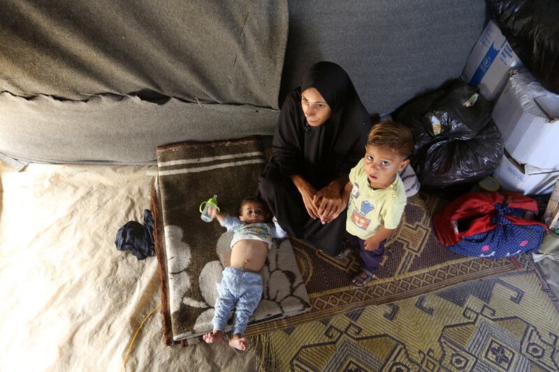 A woman sits on a rug on the ground with two little children