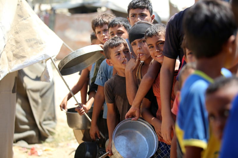 Palestinian boys, some holding pots, stand in a line at a food distribution site.