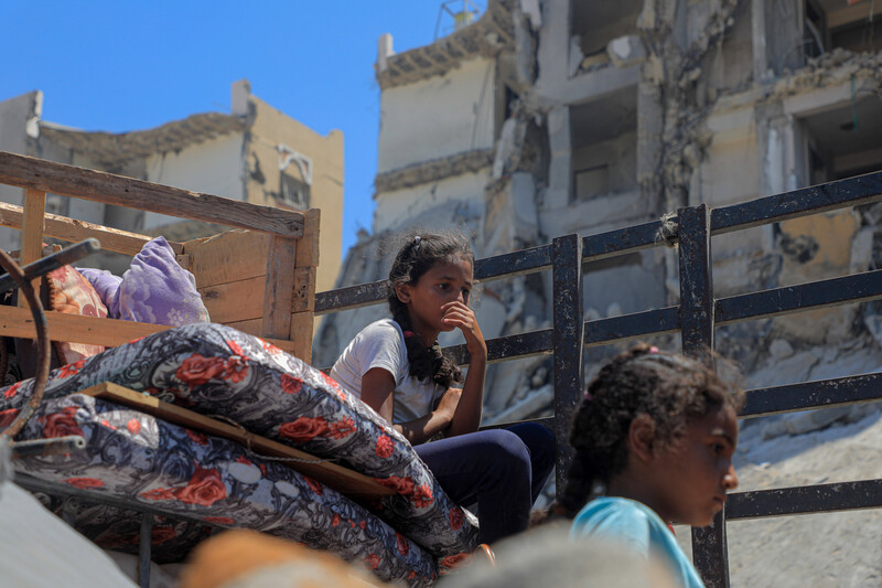 Two children sit amidst mattresses with housing destruction in background