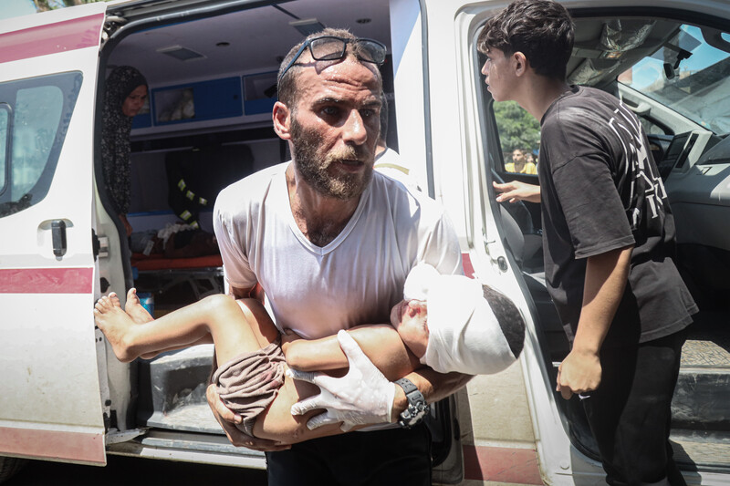 A man carries an injured child out of an ambulance with an older child and woman in the background