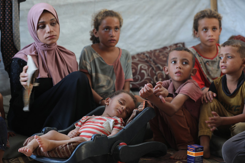 A mother and her children sit inside a white tent. A sleeping baby is laying in a carseat in the middle. 