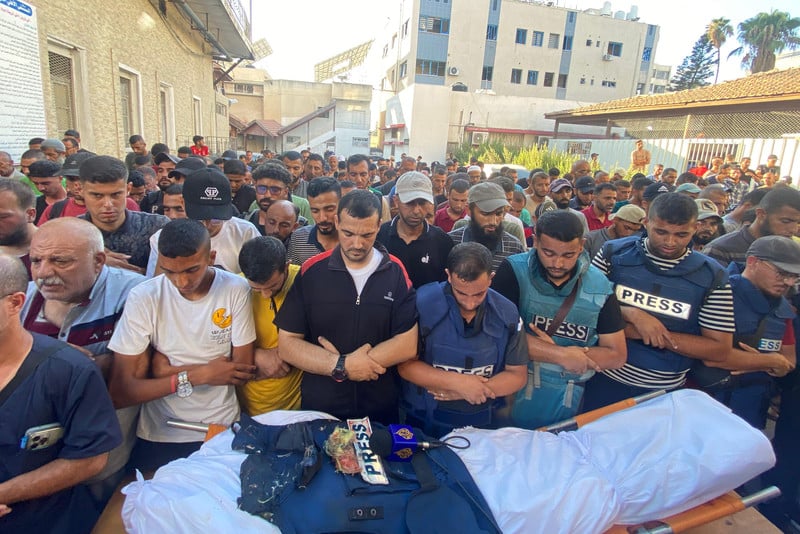 A crowd of mourners pray over a body, wrapped in a white shroud, with a damaged press jacket and an Al Jazeera microphone placed on top.