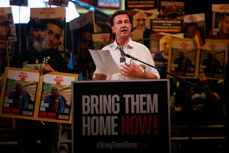 Steffen Seibert stands at a lectern surrounded by people carrying signs supporting Israelis held in Gaza