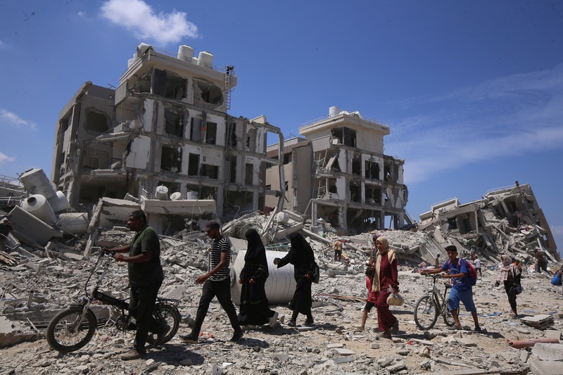 Several people, two of them holding bicycles, walk along a rubble-strewn street alongside completely bombed-out buildings