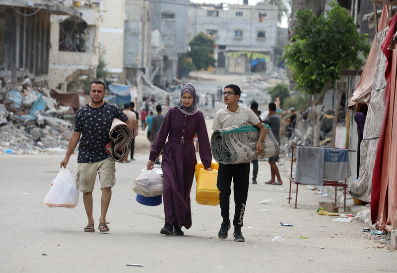 Three people walk with bags and blankets on a backdrop of ruined buildings