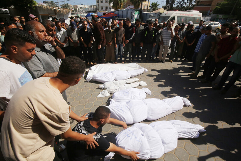 A group of mourners surround six corpses of various sizes wrapped in white shrouds