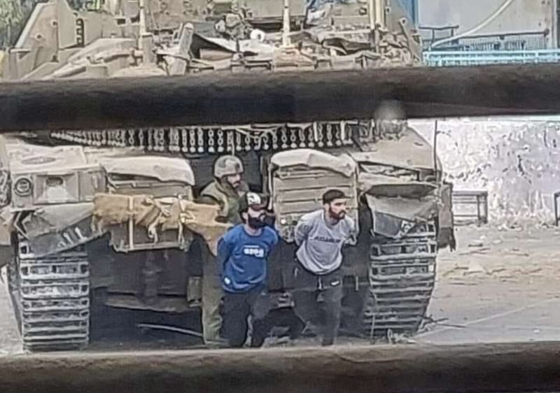 Two men with arms behind their back walk in front of an Israeli tank