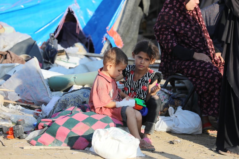 Two children sit in front of a tent that has become their shelter