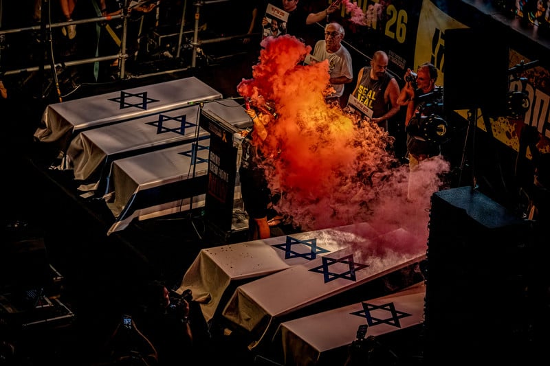 A person holds a flare emitting fiery-looking orange smoke while standing next to coffins draped in Israeli flags