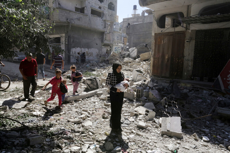 Palestinian children and adults, including a woman holding an infant, step across a rubble-strewn street