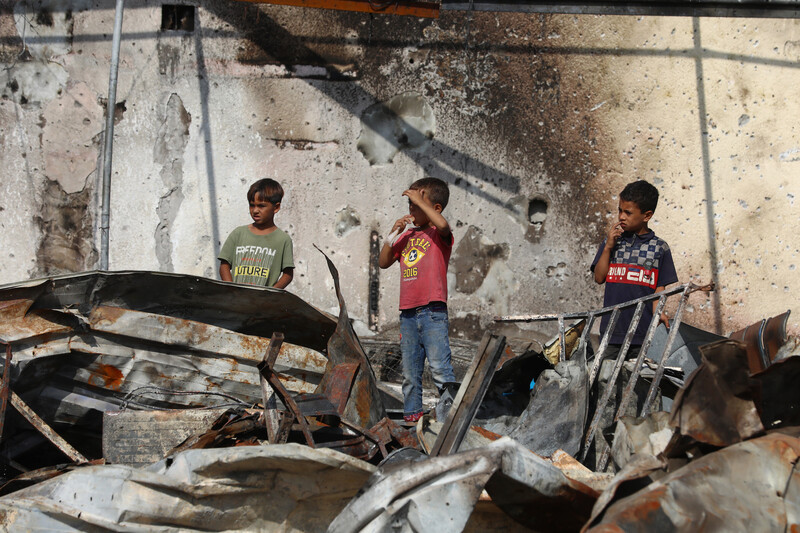 Three children stand on the charred and twisted ruins of shelters hit by an Israeli airstrike.