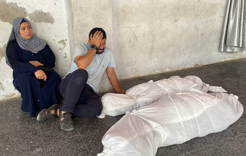 A man and a woman mourn next to two bodies wrapped in white shrouds