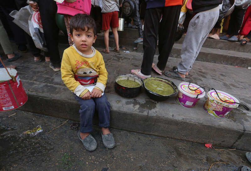 A young Palestinian child sits alongside food as others queue