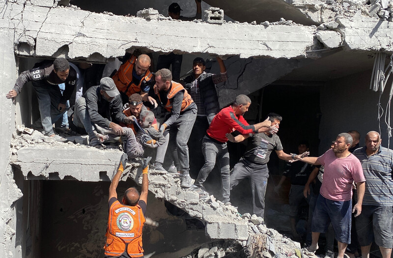 A group of people, some of them wearing orange civil defense vests, work to pull people out of the rubble of a destroyed building