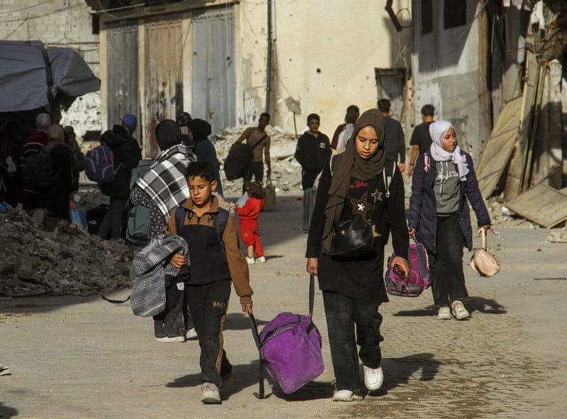 people carying their belongings walk down a street strewn with rubble