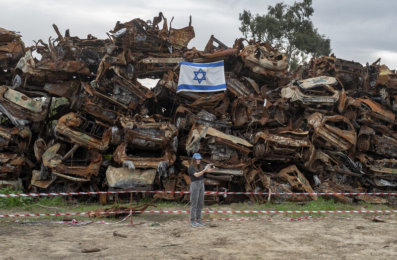 A woman takes a selfie in front of a stack of crushed cars and an Israeli flag