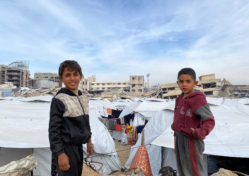 Two boys stand facing the camera in front of white tents, with destroyed buildings in the background