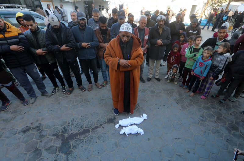 A man wearing a dark orange coat prays over two tiny bodies in white shrouds. A crowd of mourners is behind him.