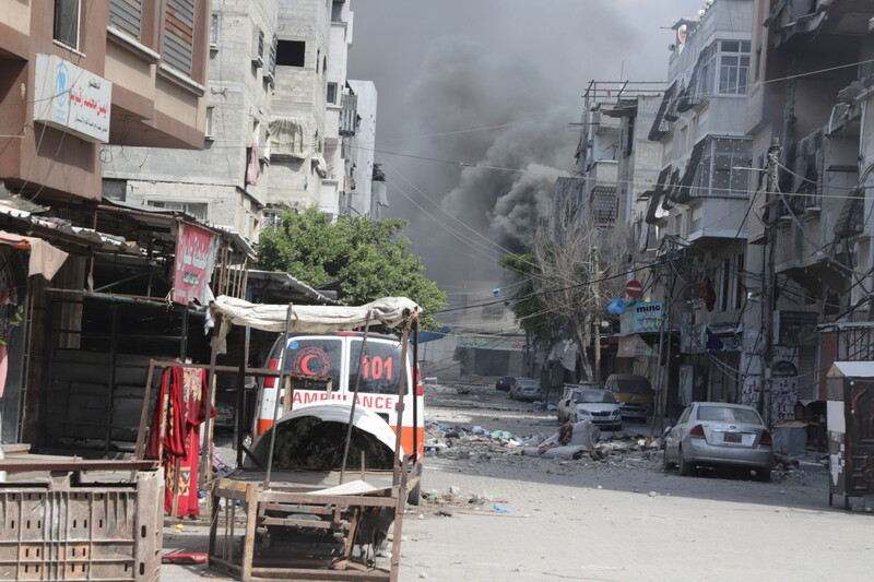 An ambulance in foreground and huge dust cloud in background caused by Israeli bombing