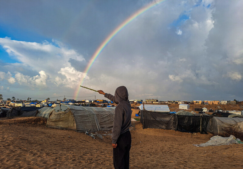 Amidst tents of displaced families, a boy points a stick at a rainbow arching across the sky. 