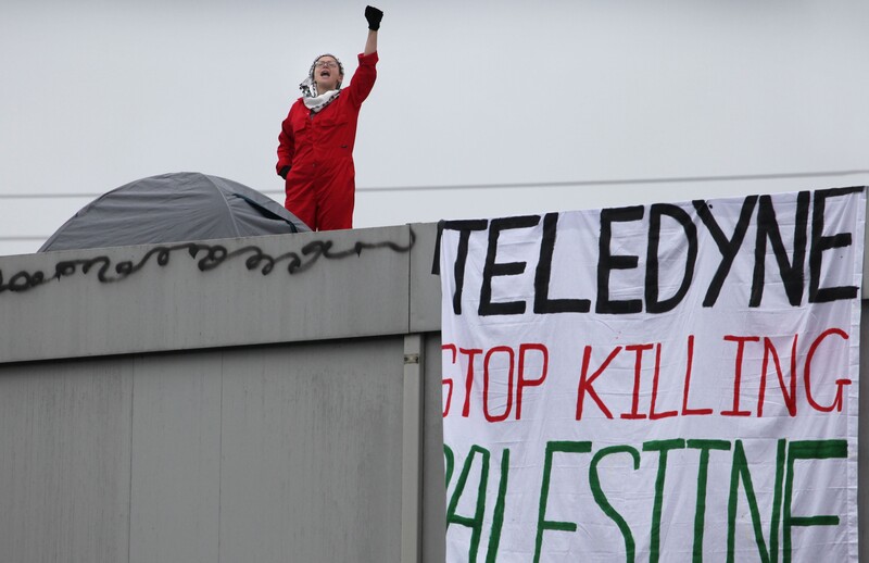 A woman in a boiler suit standing on a factory rooftop raises her fist