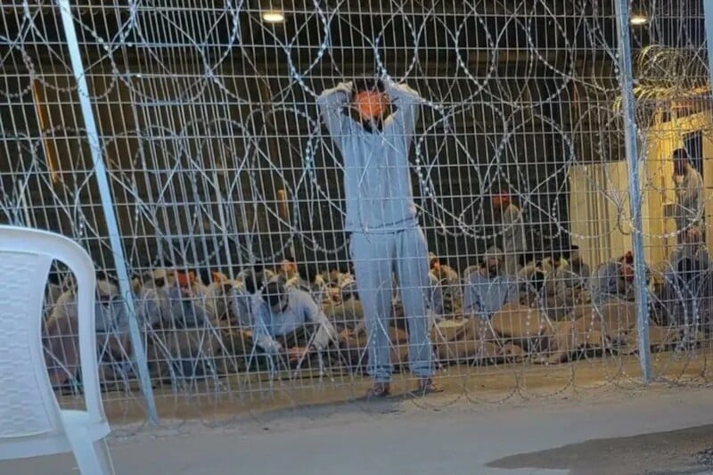 Men blindfolded and bound sit or stand behind razor wire fence