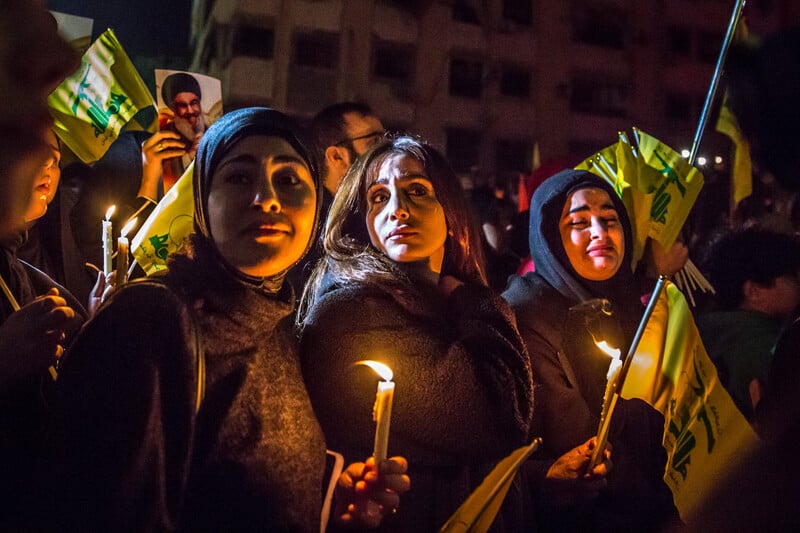 Three young women with sad expressions on their faces hold lit candles and Hizballah flags