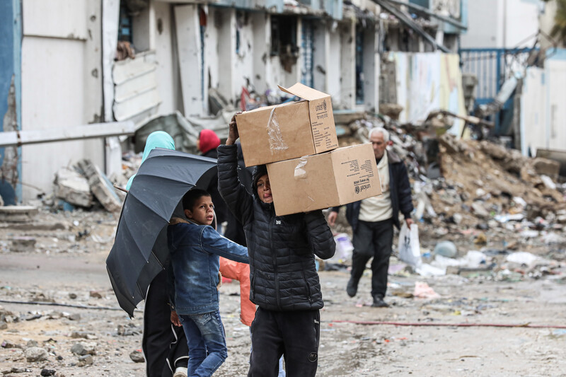 A boy holding an umbrella looks at another boy who is carrying boxes on his shoulders