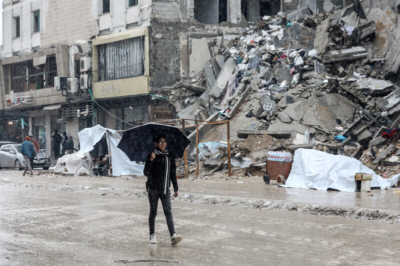 A Palestinian youth, wearing black jeans and a black sweatshirt, carries an umbrella past piles of rubble
