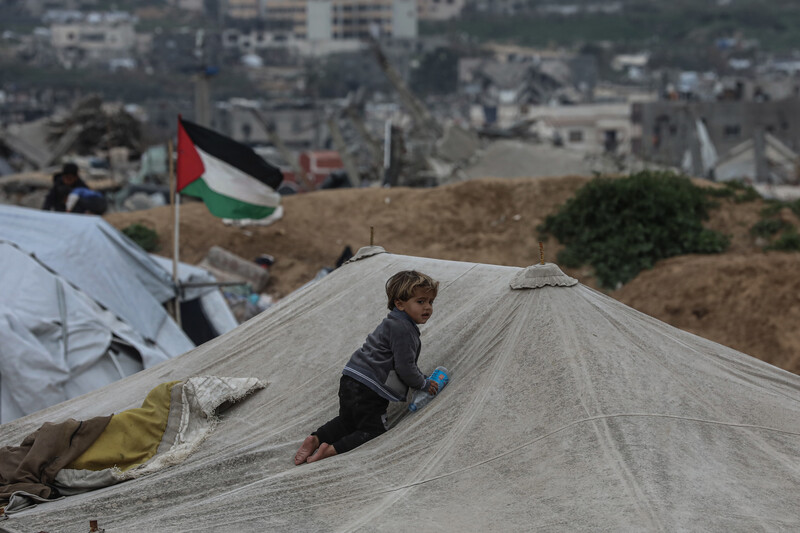 A small child climbs on the top of a white canvas tent. A Palestine flag waves behind them.