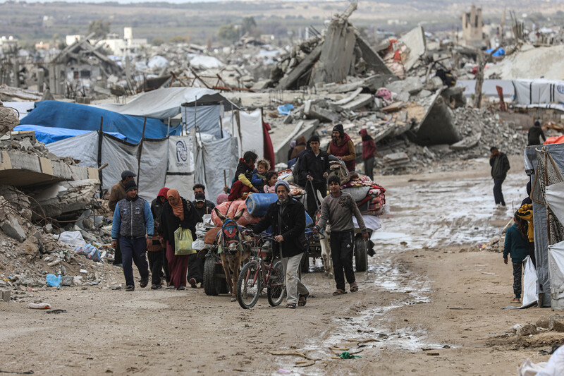 People carry their belongings on animal-drawn carts while walking along dirt road with makeshift shelters built in debris in the background