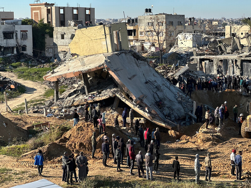 Dozens of people stand around collapsed building with crater next to it