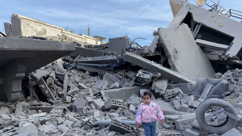 A small child stand in front of the rubble of a destroye home
