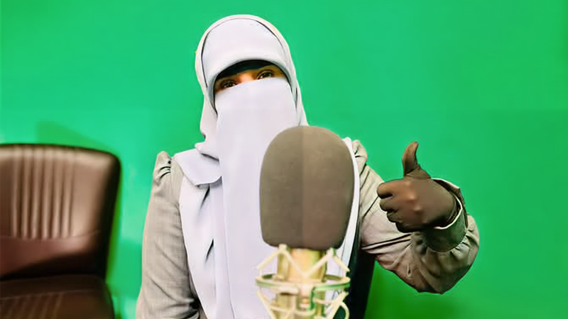 Woman wearing niqab gives a thumbs up while sitting between a microphone and a green screen