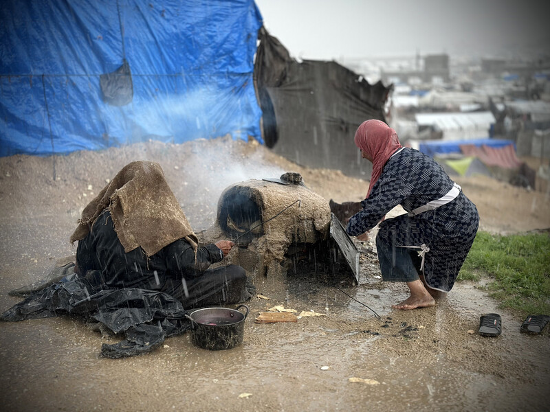 Women tend to a clay oven in rainy weather with tent encampment in background