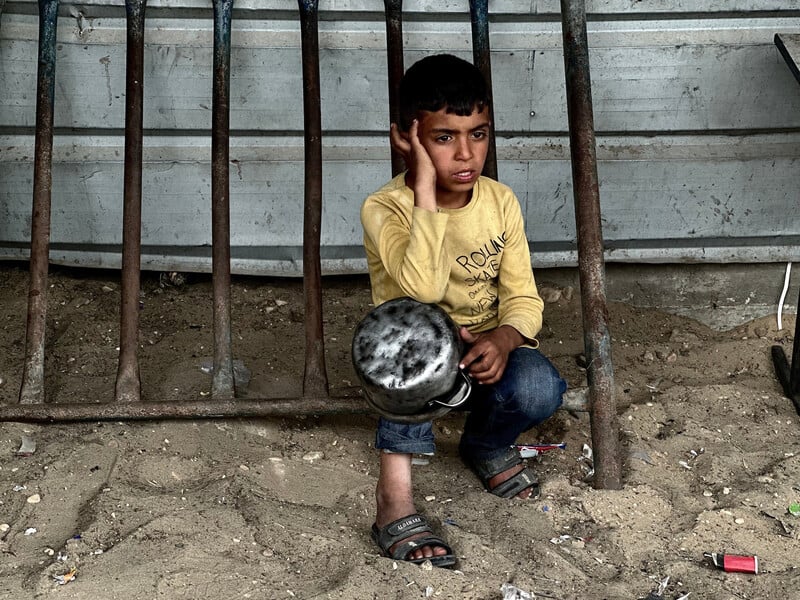 A boy in a yellow shirt kneels on the ground with an empty pot in his hands