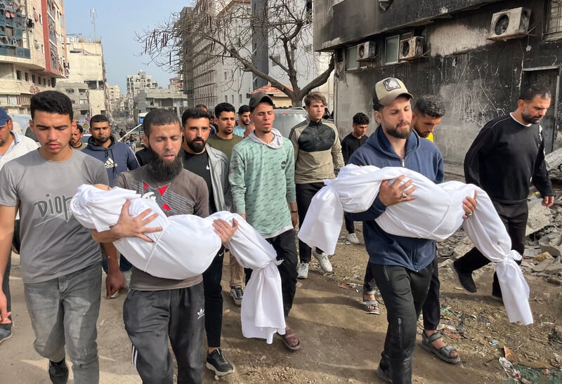 A group of men, two of them carrying small bodies wrapped in white shrouds, walk down a street