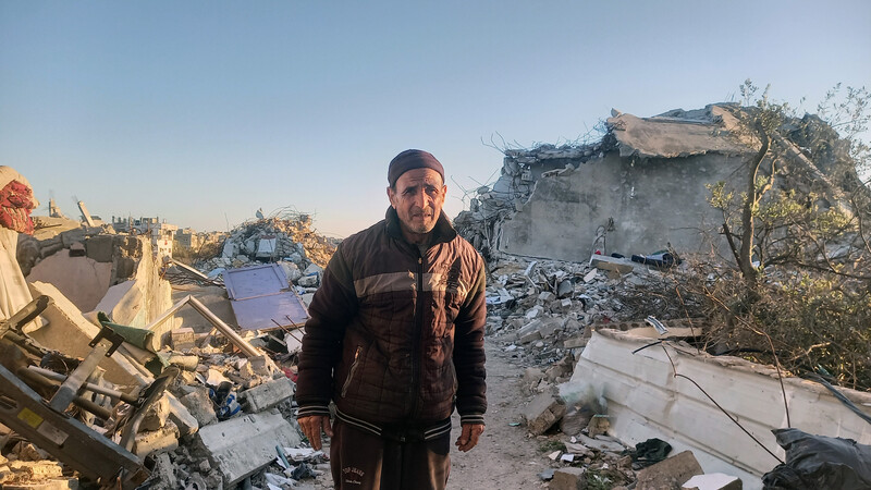 An older man looks at the camera while standing on narrow road in between destroyed buildings