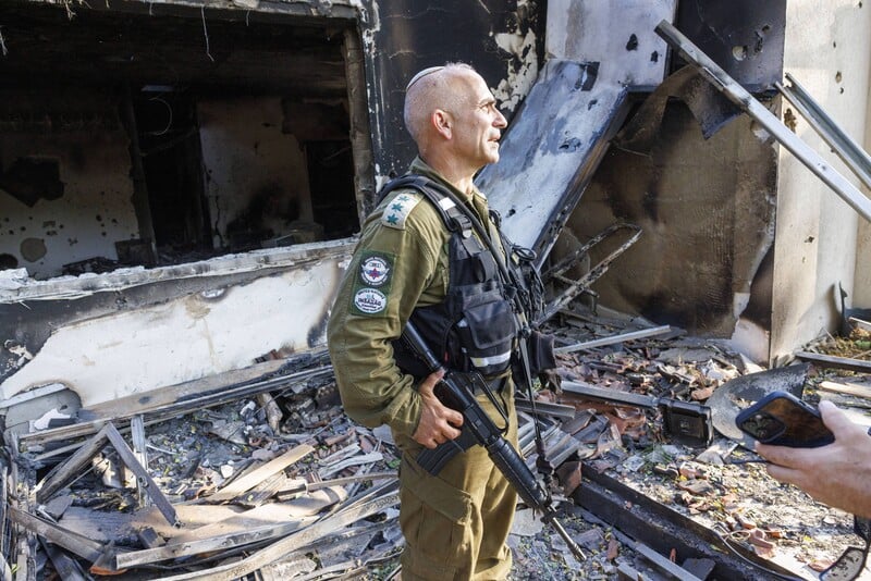 Man in military uniform stands in front of badly burned and damaged house